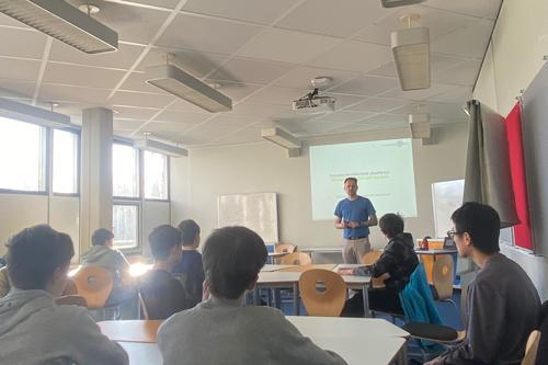 In einem Klassenraum steht ein Mann vor einer Gruppe Jungen und hält eine Präsentation. Die Schüler sitzen an Gruppentischen, im Hintergrund ist eine Folie an die Wand projiziert.