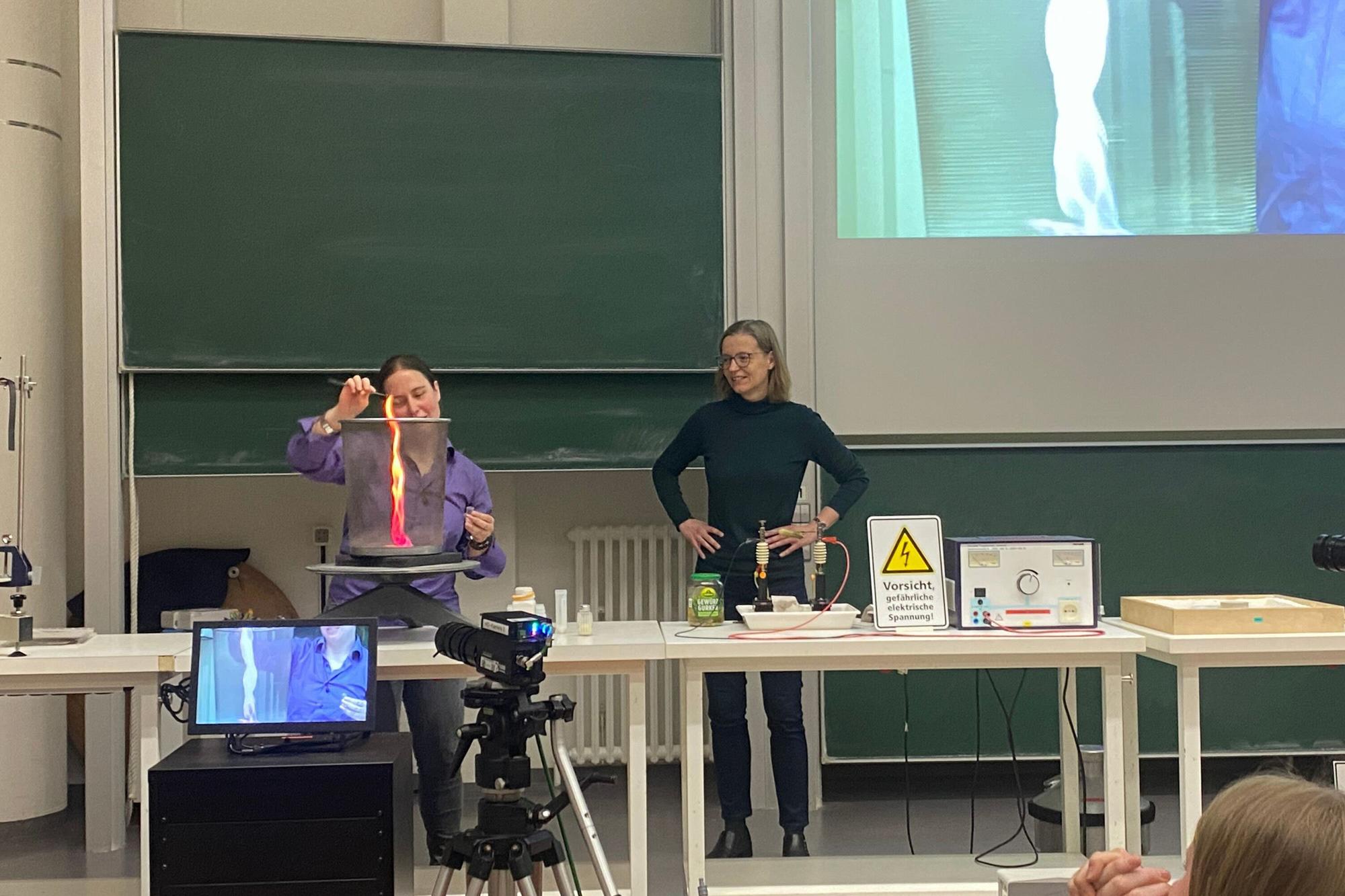 Zwei Frauen führen vor einer Tafel im Hörsaal ein physikalisches Experiment vor. Eine hält ein Reagenzglas mit leuchtender Flüssigkeit, auf dem Tisch stehen Laborgeräte und eine Kamera filmt mit.