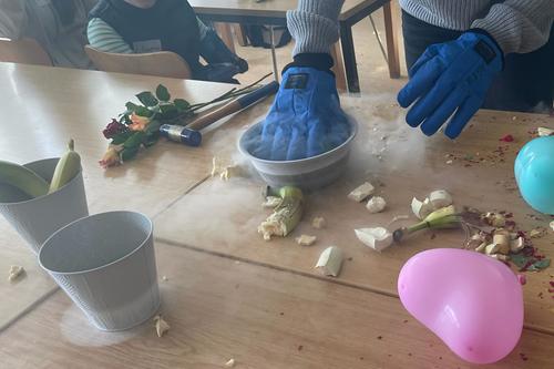Eine Person mit blauen Schutzhandschuhen steht an einem Tisch mit dampfender Schüssel. Daneben liegen zerkleinerte Bananen, Blüten, ein Hammer und bunte Ballons. Kinder beobachten das Geschehen im Hintergrund.