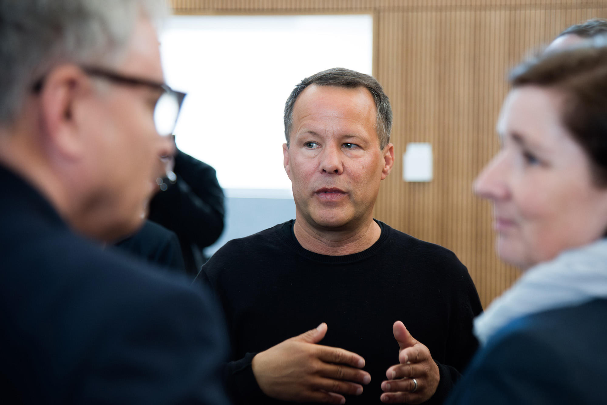 Timm John im schwarzem Pullover spricht gestikulierend mit mehreren Personen in einem modernen Raum mit Holzverkleidung. Die Szene wirkt wie ein intensives Gespräch oder eine Diskussion in einem beruflichen Umfeld.